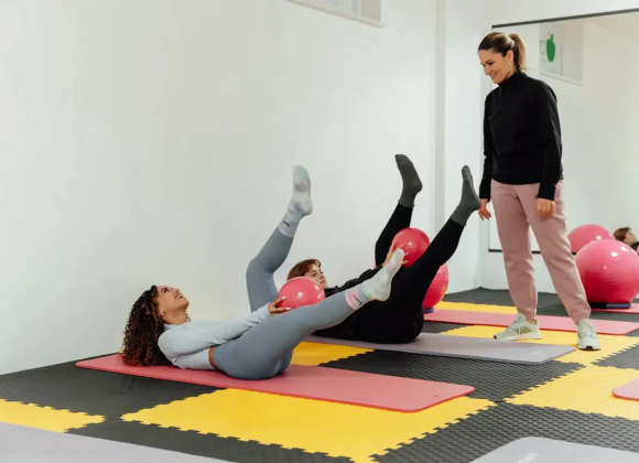 Instructor coaching two women through a core strengthening exercise on mats, each holding a small exercise ball